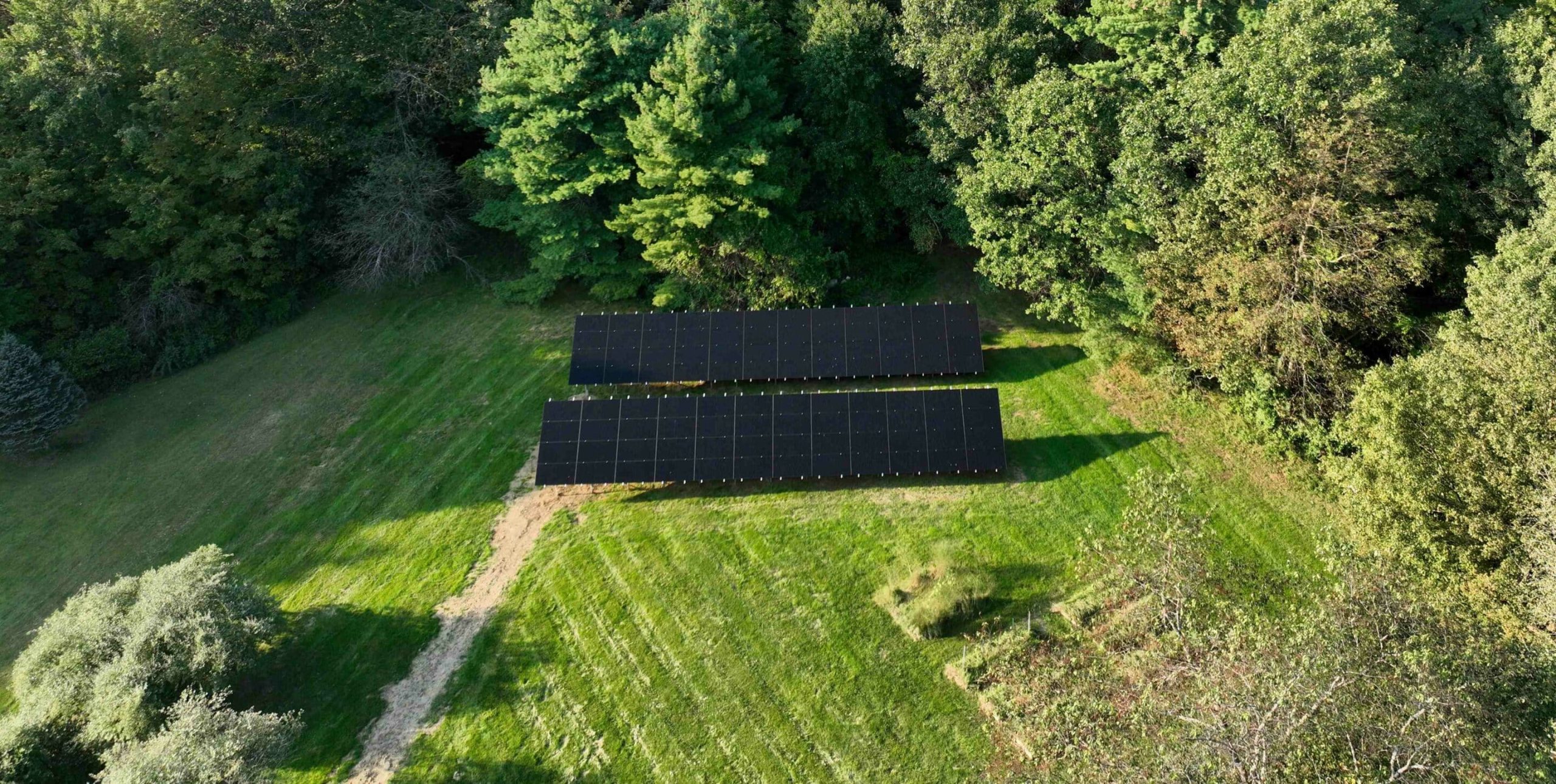 Aerial view of two rows of solar panels on a grassy field bordered by dense green trees.