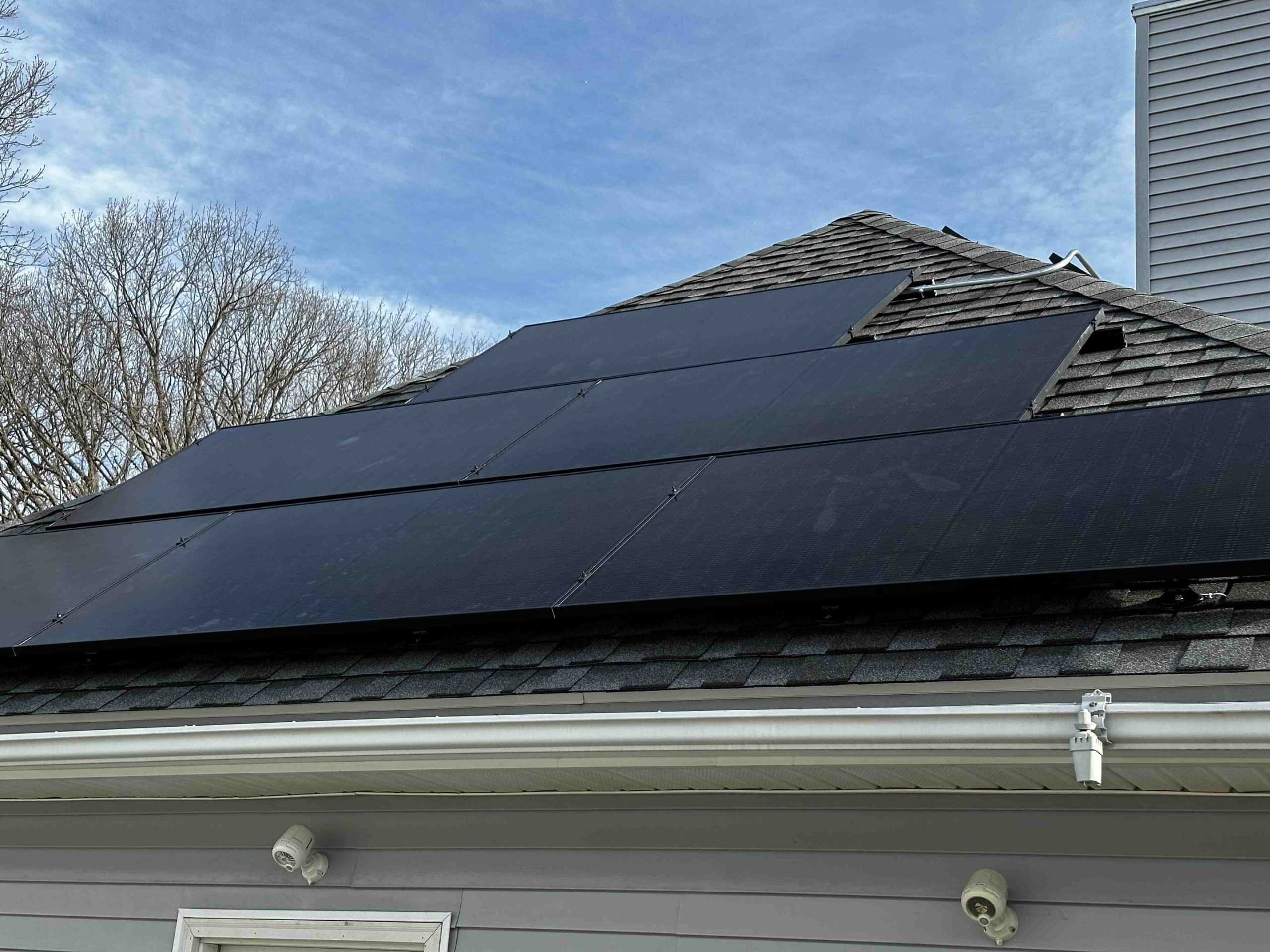 Solar panels installed on the roof of a house with a clear sky and leafless trees visible in the background.