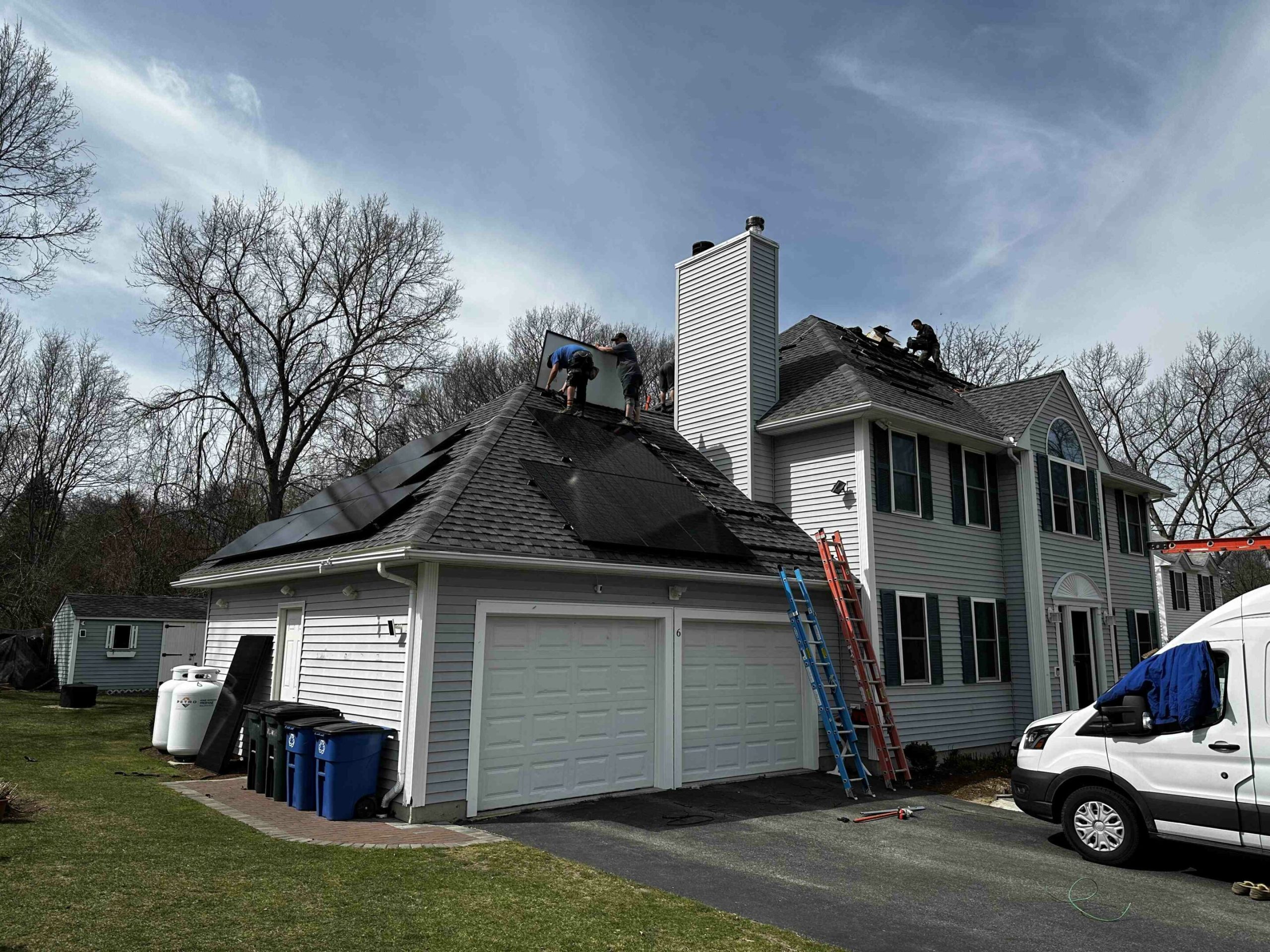 Workers install solar panels on the roof of a two-story house with a driveway, white van, and trees in the background.