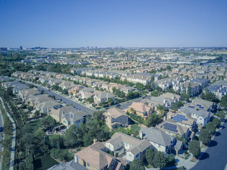 Aerial view of a suburban neighborhood with rows of houses, tree-lined streets, and a cityscape visible in the distant background under a clear sky.