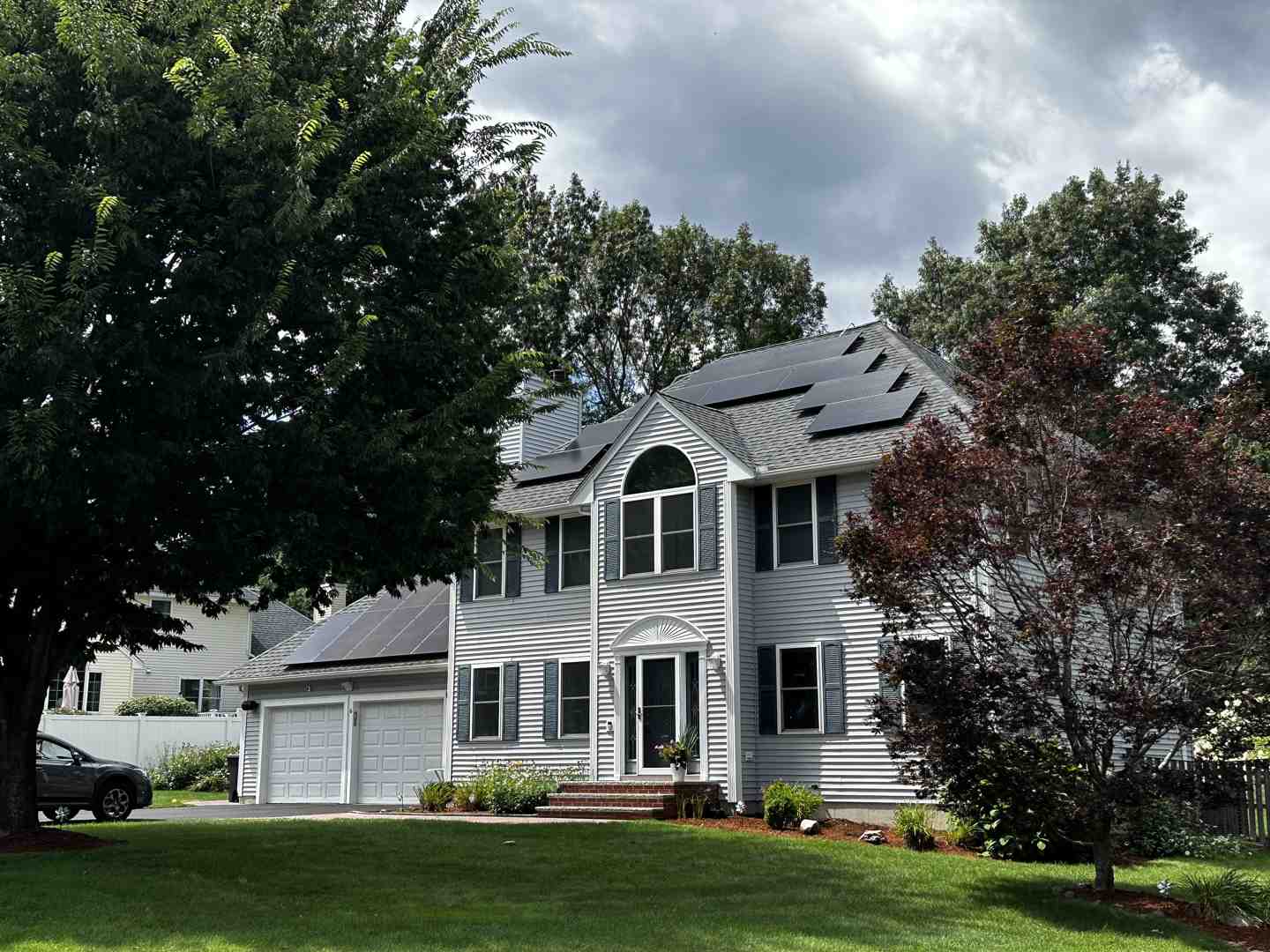 A two-story gray house with solar panels on the roof, surrounded by trees and a well-kept lawn under a partly cloudy sky.