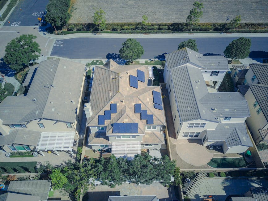 Aerial view of suburban houses, with the central house featuring solar panels on its roof and surrounded by neighboring homes and trees.