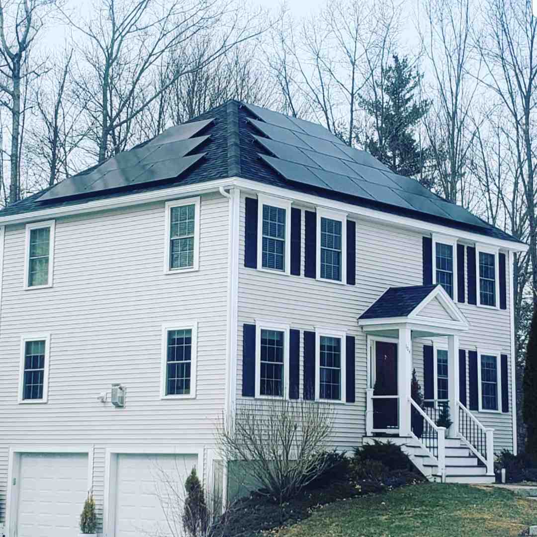 Two-story white house with black shutters and solar panels installed on the roof, surrounded by leafless trees and a small front yard.