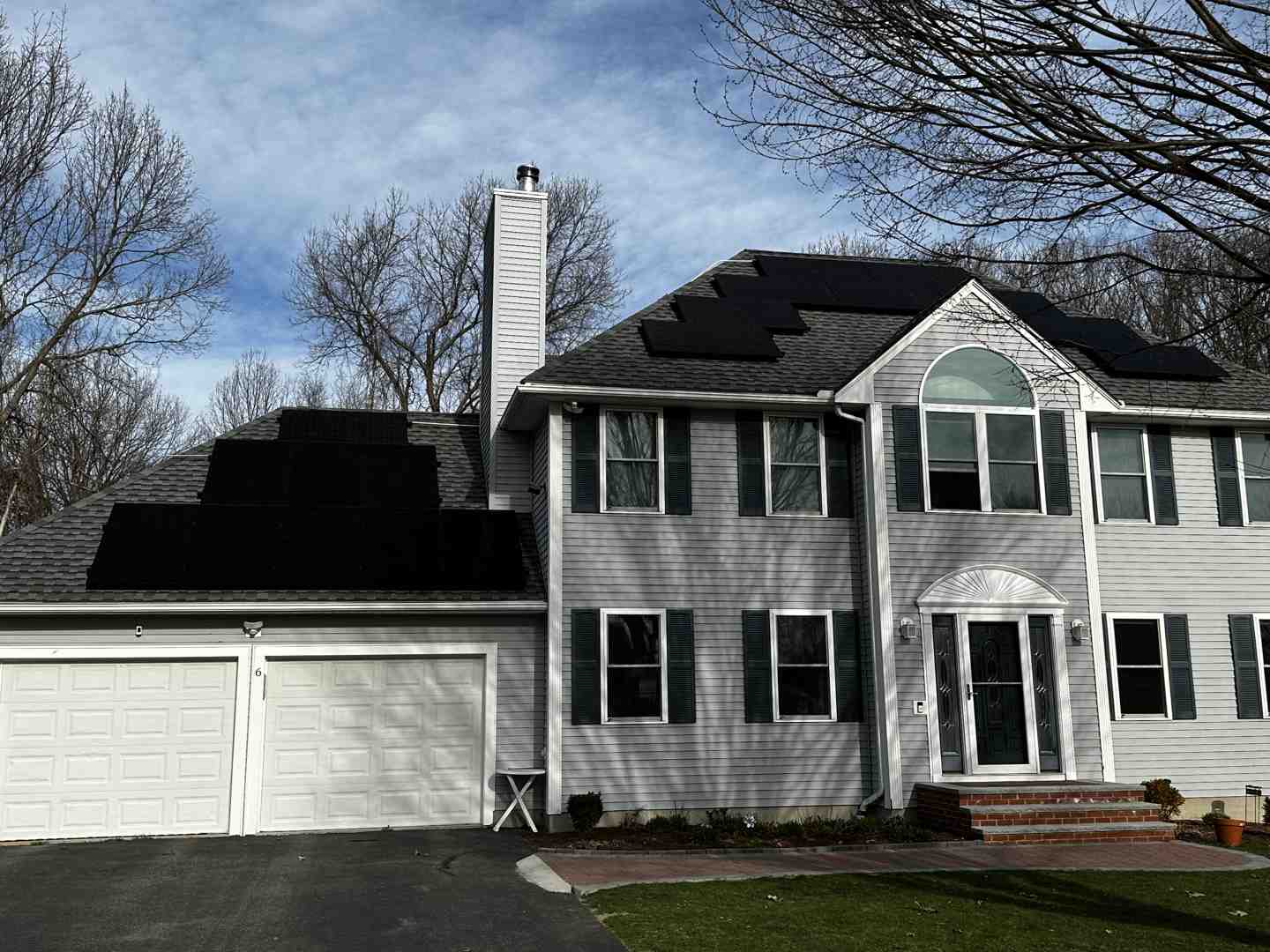 White two-story house with solar panels on the roof, double garage, and bare trees in the background on a cloudy day.