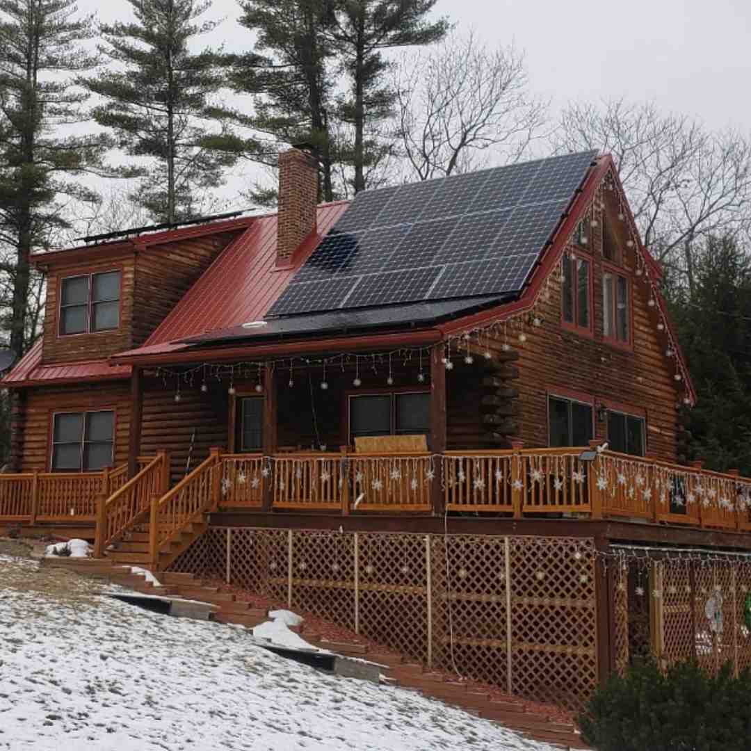 A log cabin with solar panels on the roof, a red metal roof, wooden deck, and patches of snow on the ground surrounded by pine trees.