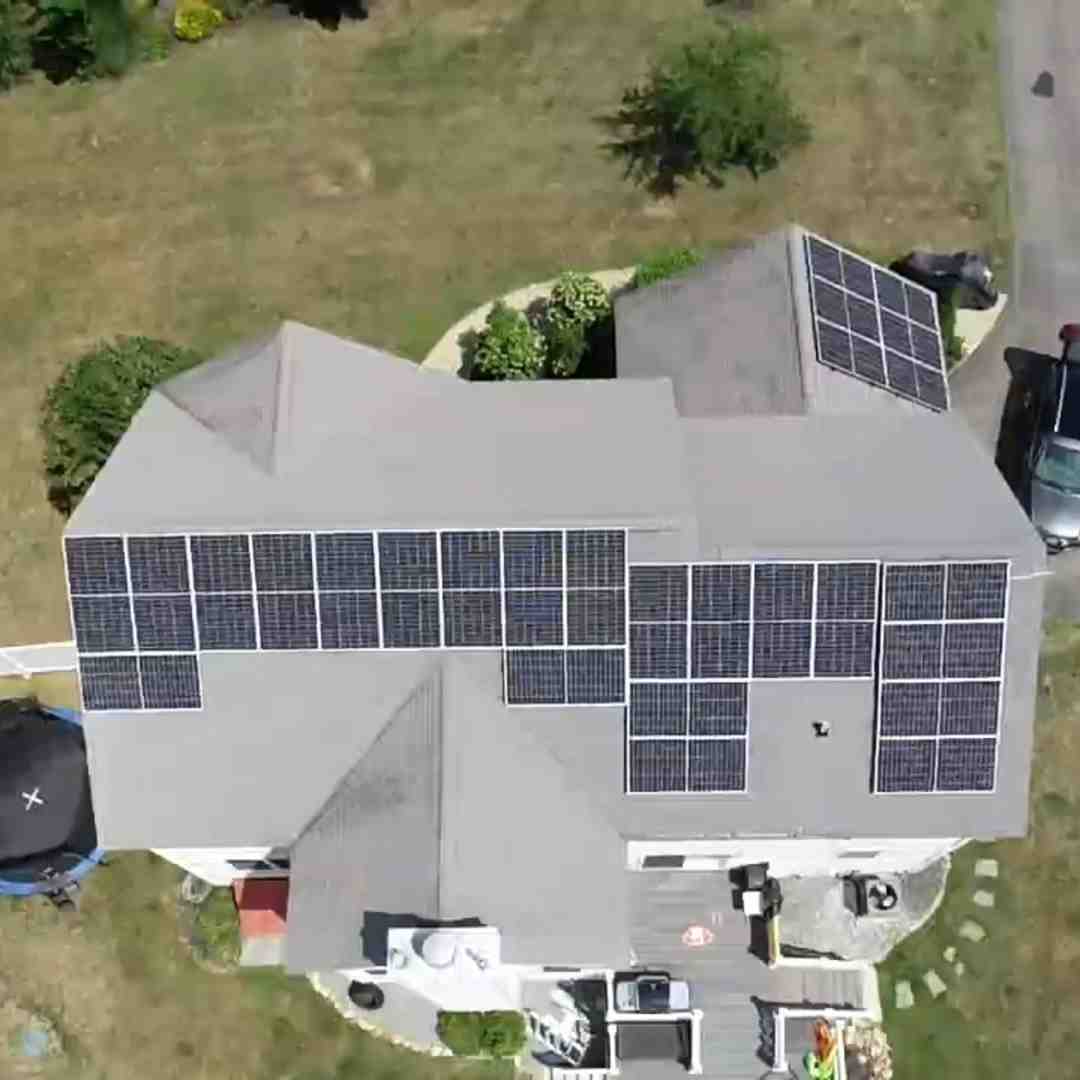 Aerial view of a house with multiple solar panels installed on its roof and a few more on a nearby structure, surrounded by grass and a driveway.