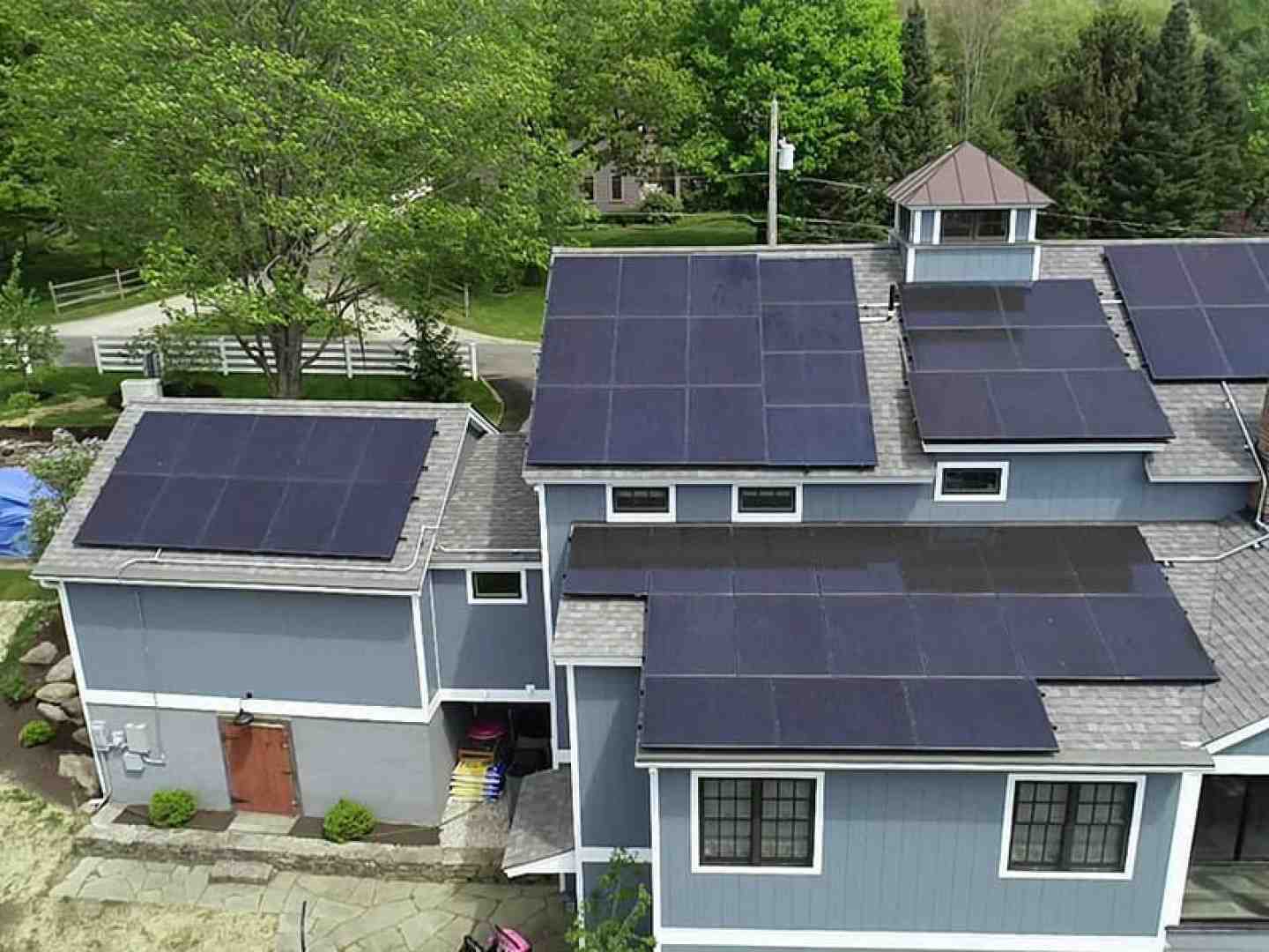 Aerial view of a gray house with multiple black solar panels installed on its roofs, surrounded by green trees and a yard.