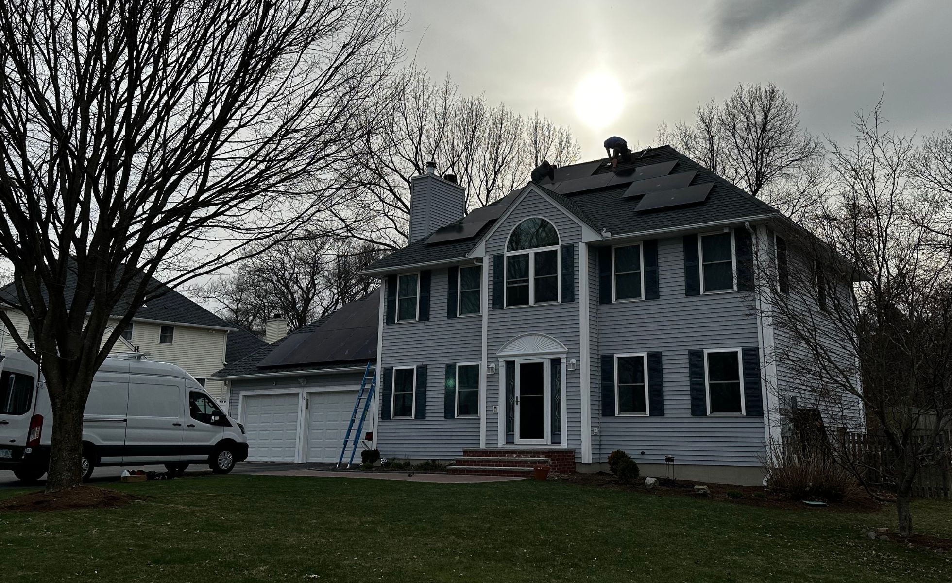 An installer from a local Solar Power Company fits solar panels on a two-story house; a white van and ladder are nearby under cloudy skies.