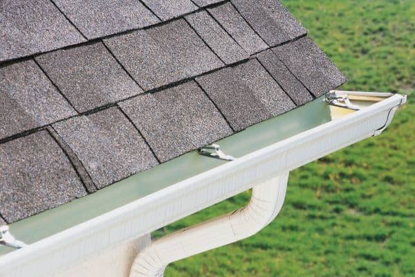 Close-up of a house roof with gray asphalt shingles and pristine white gutter installations, set against a lush green grass background.