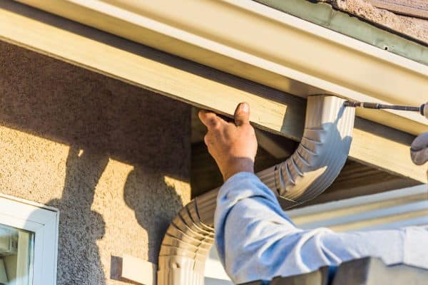 A person performs gutter installations by securing a metal rain gutter to the house roof edge with a drill, ensuring it’s properly in place.