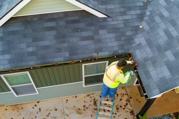 A person in a safety vest stands on a ladder cleaning leaves from the gutters of a house with a gray roof, preparing for gutter installations.
