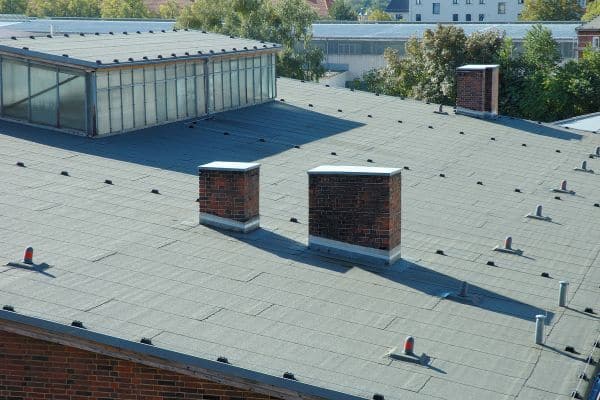 Flat rooftop with two brick chimneys, ventilation fixtures, and a glass structure—ideal for Commercial Roofing Services—surrounded by trees and buildings.